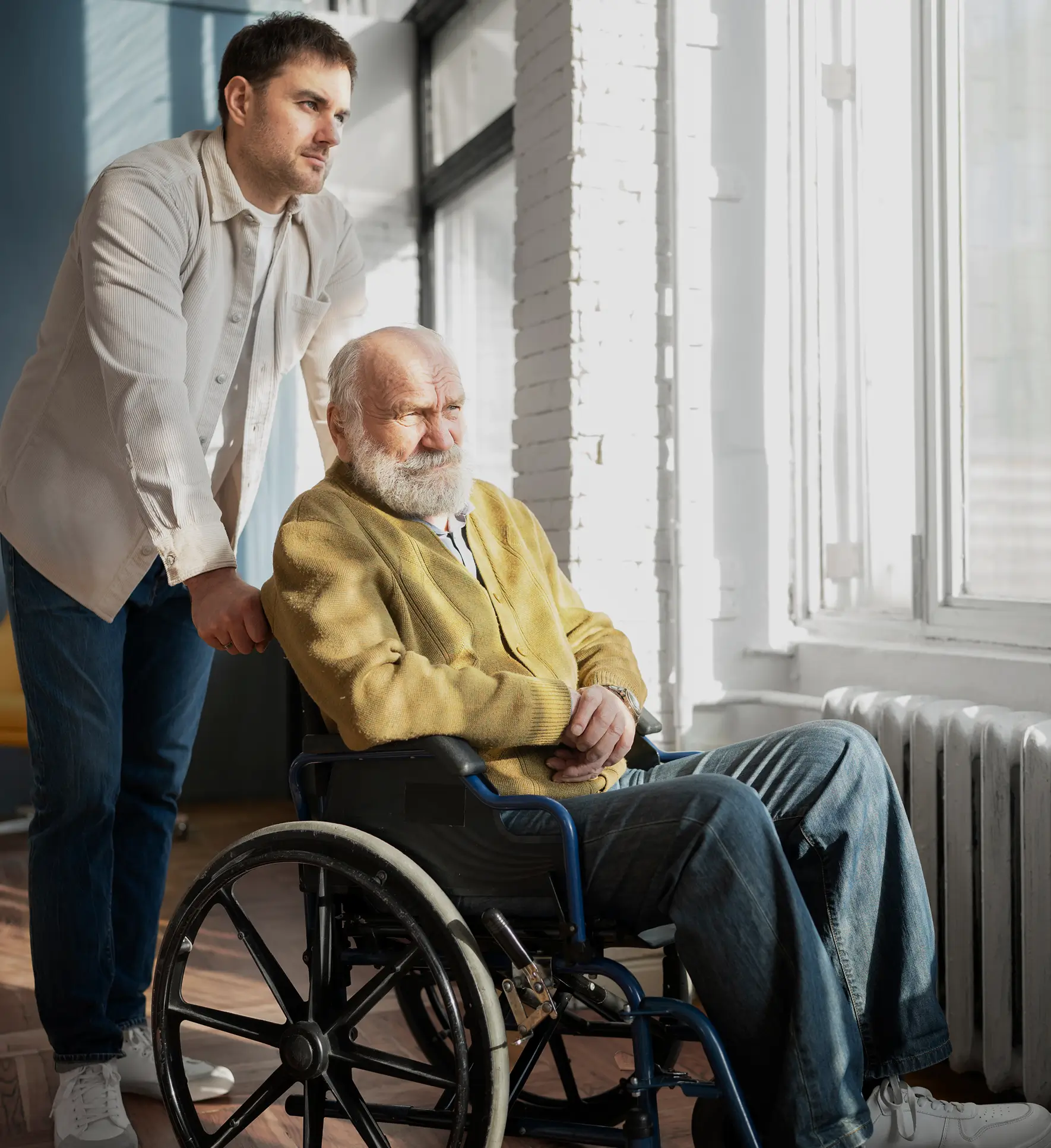 A man helping an older man in his wheelchair by a window at home